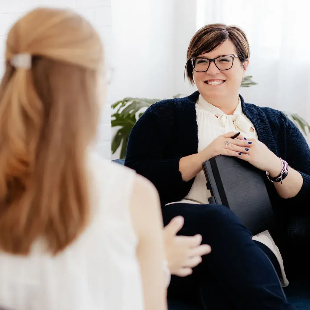 A smiling woman with glasses holding a notebook, seated across from a person with long hair in a bright room with plants