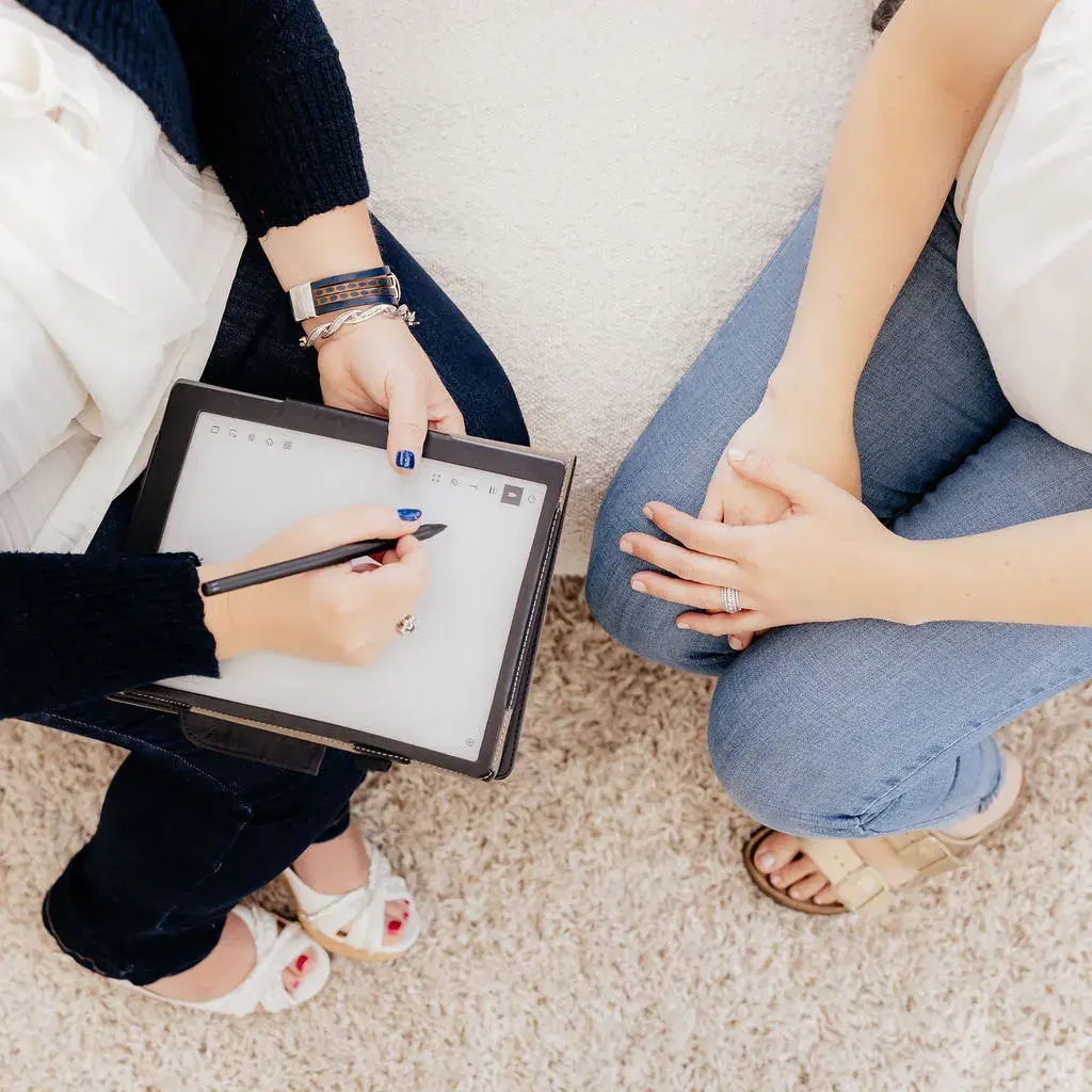 Overhead view of two seated people, one writing on a tablet with a stylus and the other resting her hands on her knee