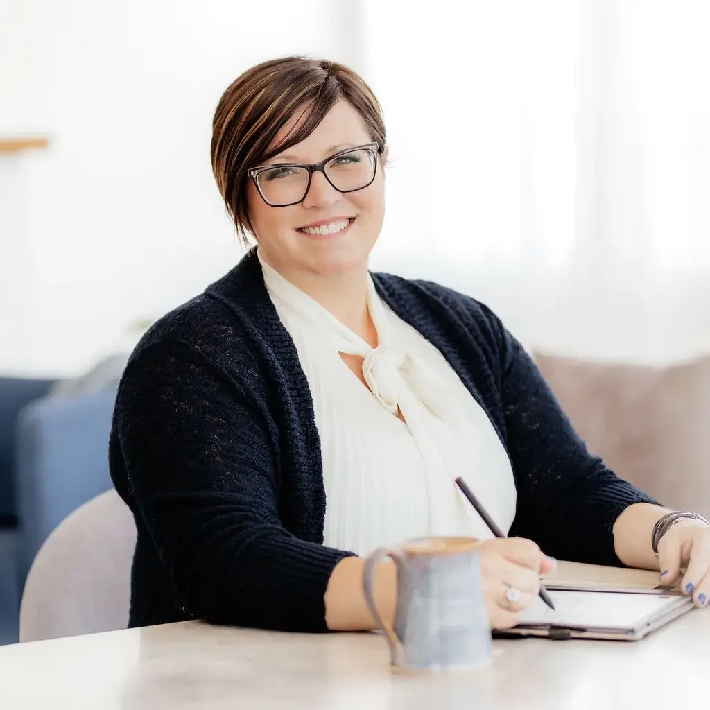 A smiling woman with glasses seated at a table with a tablet, a stylus and a ceramic mug
